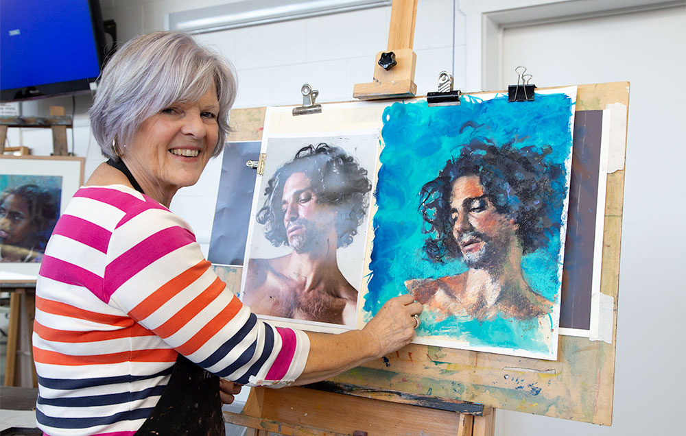 Liz Turner working on a portrait of a young man with long, dark curly hair created in acrylic ink and pastel.